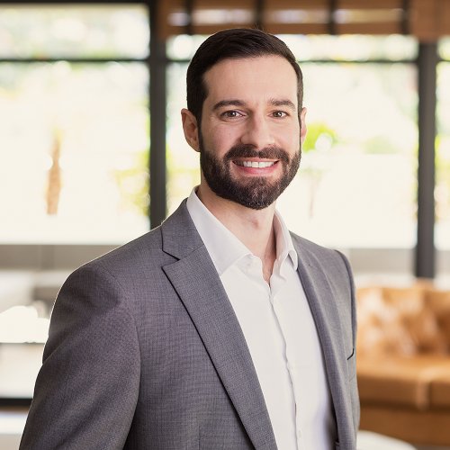 Homem sorridente de cabelo e barba escuros, vestindo terno cinza e camisa branca, em um ambiente interno iluminado.